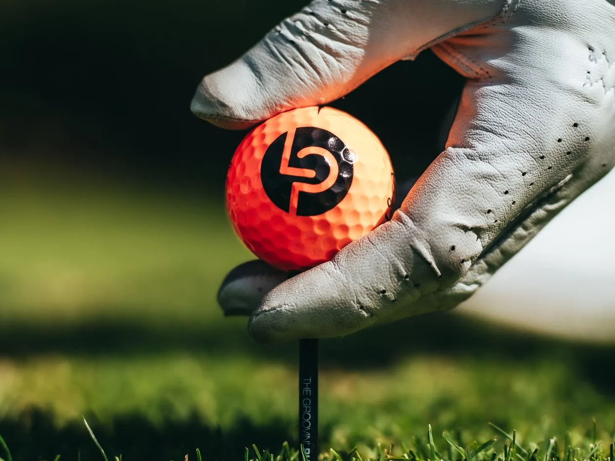 Behandschuhte Hand legt einen orangefarbenen LP-Golfball aufs Tee